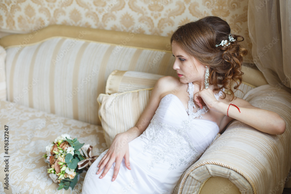 Beautiful young Bride with classic hairstyle sits on a sofa in a bedroom. Classic fish-silhouette white wedding dress. Wedding fashion Beautiful young Bride with classic hairstyle sits on a sofa in a bedroom. Classic fish-silhouette white wedding dress. Wedding fashion