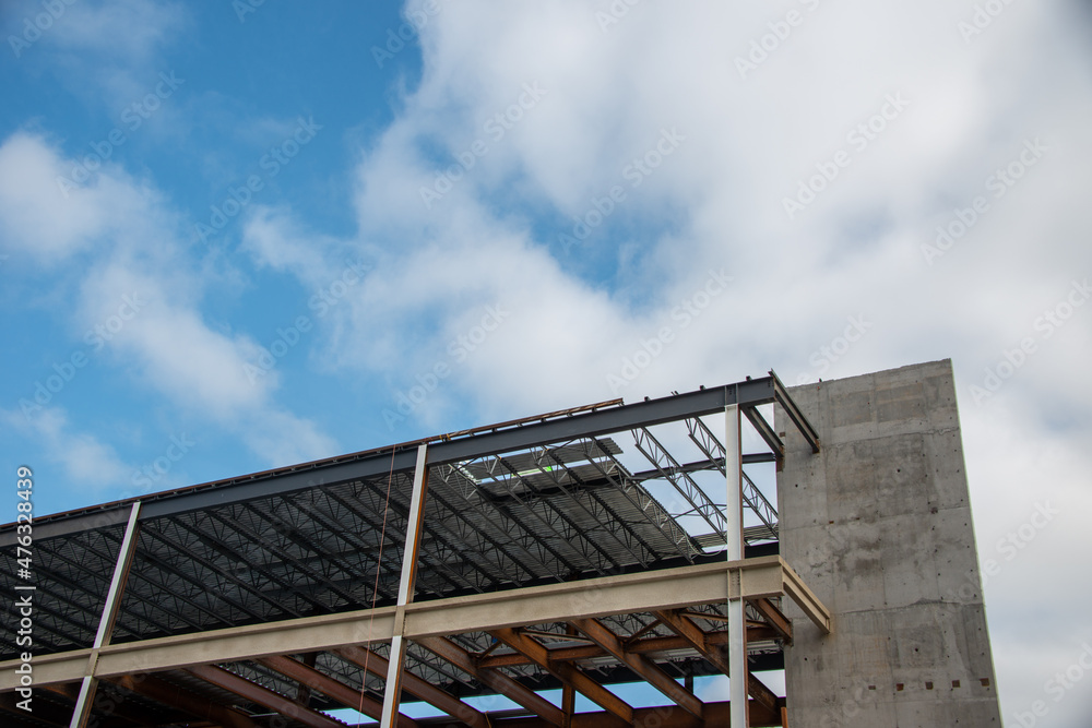 A large multiple story metal building wall with steel beams against a blue sky. The industrial structure in the corner of a skyscraper building with prefabricated engineering formwork.