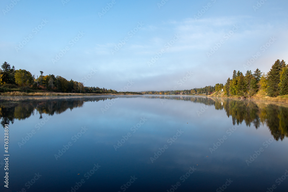 A wide river with smooth blue water. There are luscious green trees along the riverbank. The sky is clear blue. The calm and peaceful pond water is reflecting the sky and trees.