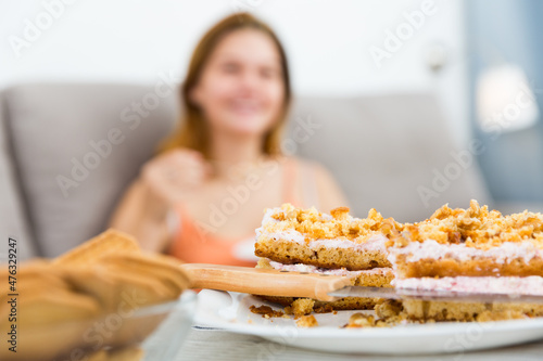 Young smiling female eating sweet cake on sofa with spoon indoors.