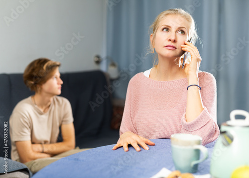 Woman having serious phone conversation while sitting at table at home against background of her upset teenage son photo