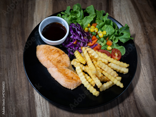 Grilled salmon steak with vegetable and french fries on plate, on wood table, top view.