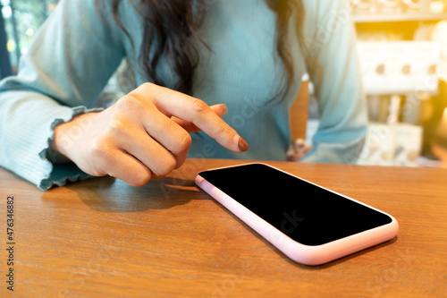 Young beautiful Asian woman with green color shirt searching information on her smart phone. Close-up woman's finger points at a mobile phone screen on the wooden table at cafe and restaurant.