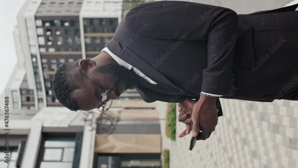 Vertical tracking of African-American businessman in suit and glasses standing on street and using mobile phone Vertical tracking of African-American businessman in suit and glasses standing on street and using mobile phone