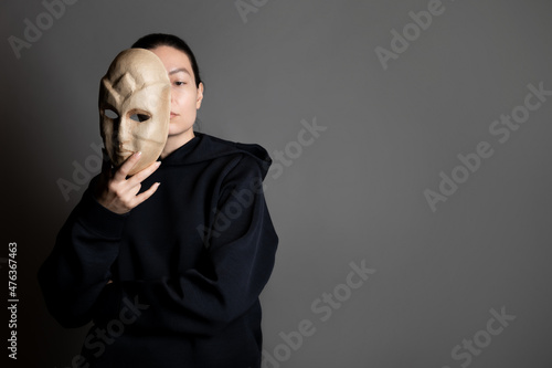 Obraz na płótnie Hiding behind a mask, a young woman in a dark hoodie hides her face with a mask, self-identification problems and impostor syndrome