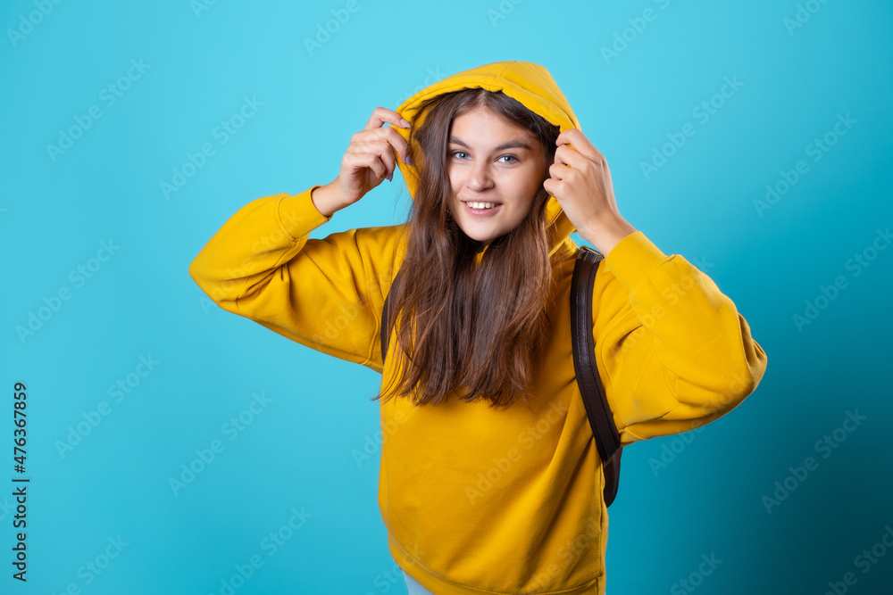 Beautiful young brunette with a hood on her head, youth oversize clothing style, portrait in a yellow hoodie on a blue background, bright colors Beautiful young brunette with a hood on her head, youth oversize clothing style, portrait in a yellow hoodie on a blue background, bright colors