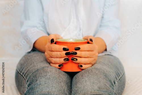 Close-up of woman holding red mug of hot tea or coffee while sitting on sofa indoors, front view. Selective focus in foreground