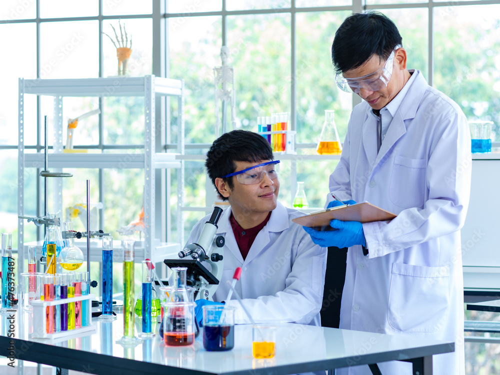 Asian young university male scientist student in white lab coat safety goggles and rubber gloves using microscope magnifying science sample in laboratory while teacher holding clipboard rating score Asian young university male scientist student in white lab coat safety goggles and rubber gloves using microscope magnifying science sample in laboratory while teacher holding clipboard rating score