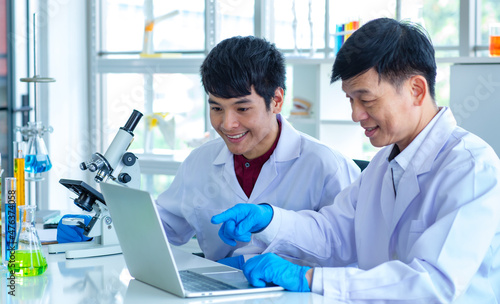 Asian professional mature male scientist teacher in white lab coat and rubber gloves sit smiling teaching science student using microscope and record analysis data in laptop computer in laboratory
