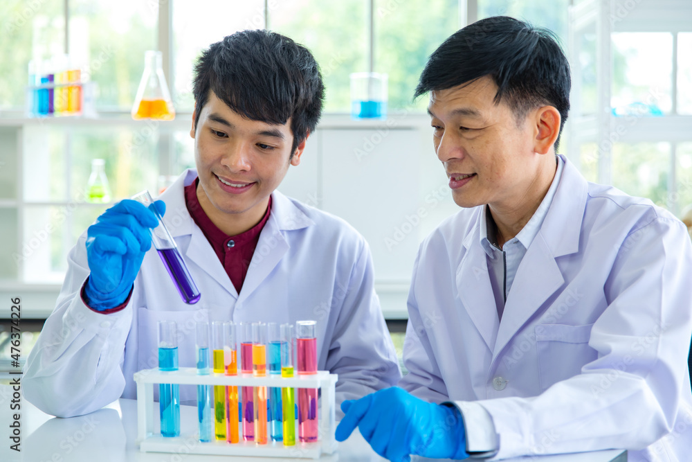 Closeup shot of scientist student hand in blue rubber gloves holding yellow solvent in glass test tube from colorful sample rack on table in laboratory while professor teacher sit smiling together