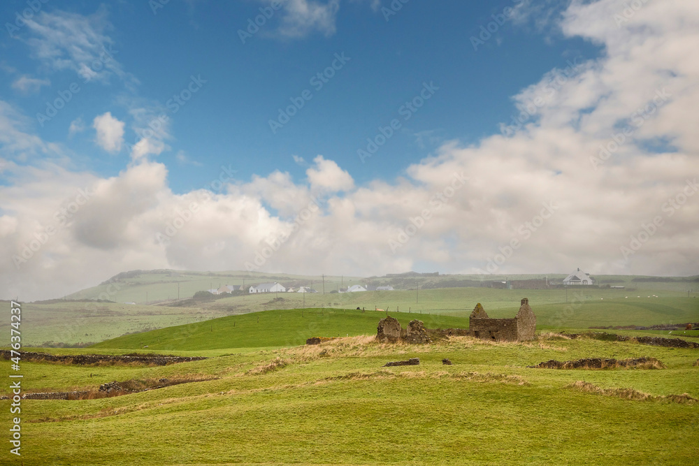 Fototapeta premium Beautiful rural landscape scene. Small stone abandoned house on a green grass hill, mountains and houses in the background. Cloudy sky. West of Ireland. Calm and peaceful mood.