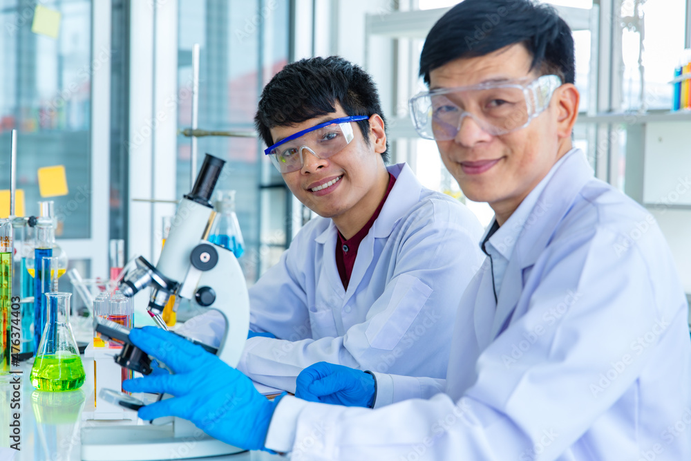 Portrait closeup shot of Asian mature male scientist teacher in white lab coat safety goggles and rubber gloves using microscope smiling look at camera with university student in blurred background Portrait closeup shot of Asian mature male scientist teacher in white lab coat safety goggles and rubber gloves using microscope smiling look at camera with university student in blurred background