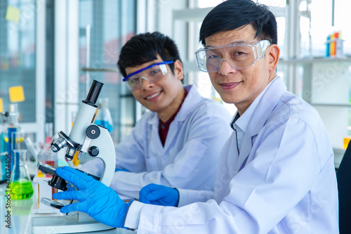 Portrait closeup shot of Asian mature male scientist teacher in white lab coat safety goggles and rubber gloves using microscope smiling look at camera with university student in blurred background