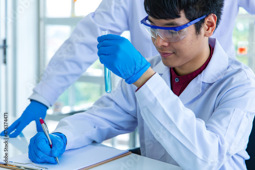 Closeup shot hands of Asian male scientist in white lab coat safety goggles and rubber gloves in blurred background careful holding pouring green sample from test tube to glass vial in laboratory photo