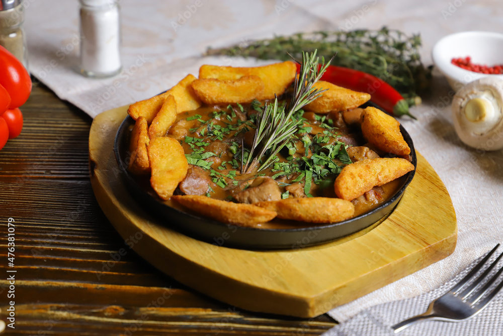 In a frying pan, fried potatoes frame pieces of meat stewed in sauce, cutlery, red bell peppers, champignons, sauce, hot peppers, spices, rosemary branch are on the table. In a frying pan, fried potatoes frame pieces of meat stewed in sauce, cutlery, red bell peppers, champignons, sauce, hot peppers, spices, rosemary branch are on the table.
