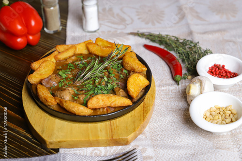 In a frying pan, fried potatoes frame pieces of meat stewed in sauce, cutlery, red bell peppers, champignons, sauce, hot peppers, spices, rosemary branch are on the table. 