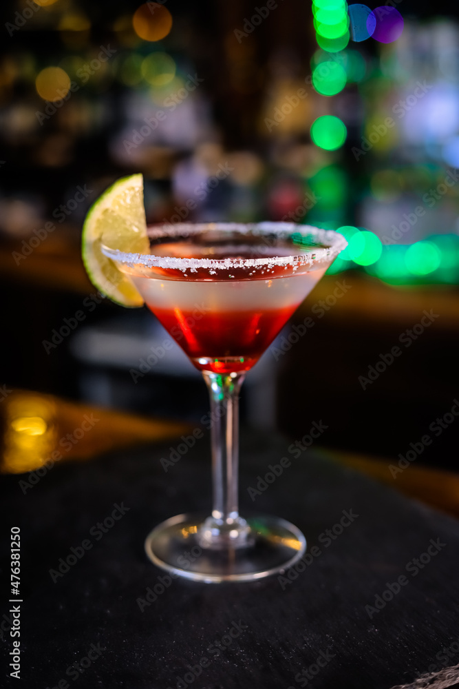 In a funnel-shaped glass on a long stem, a two-tone cocktail, dark red below, translucent white above, a lime slice on the edge of the glass. Filming of the bar menu against the background In a funnel-shaped glass on a long stem, a two-tone cocktail, dark red below, translucent white above, a lime slice on the edge of the glass. Filming of the bar menu against the background
