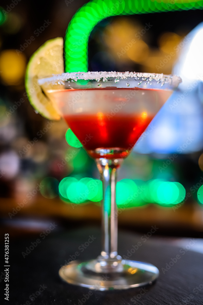 In a funnel-shaped glass on a long stem, a two-tone cocktail, dark red below, translucent white above, a lime slice on the edge of the glass. Filming of the bar menu against the background