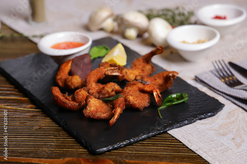 Shrimp is served with sauce. Tempura king prawns, sliced lemon, green basil leaves are served on a black stone stand. Cutlery, hot pepper pods, spices in bowls, white mushrooms.