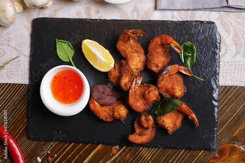 Shrimp is served with sauce. Tempura king prawns, sliced lemon, green basil leaves are served on a black stone stand. Cutlery, hot pepper pods, spices in bowls, white mushrooms.