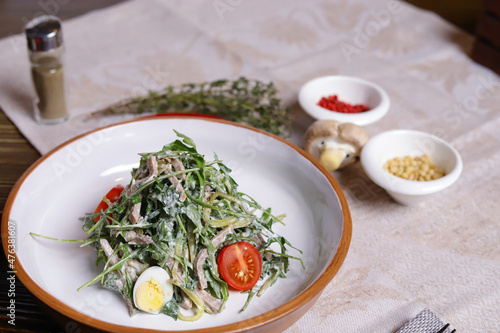 On a dark countertop, a light linen napkin, on a cutting board Served salad with rucola leaves, boiled tongue cut into long slices with sauce A couple of salt and pepper, cutlery, red peppers decorate