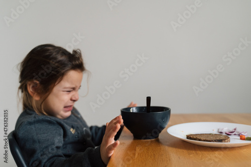 Unhappy child girl eats soup from black bowl with bread and onion. Lifestyle photo of kid in kitchen having a meal, Screaming kid. Picky eater photo