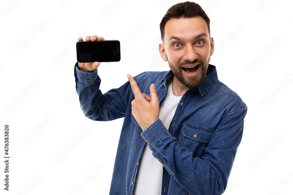 handsome young man points finger at smartphone screen with with charismatic emotions on face on white background handsome young man points finger at smartphone screen with with charismatic emotions on face on white background
