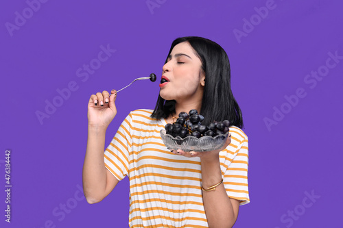 foodie girl holding bowl of grapes and grape her mouth  over purple background indian pakistani modle photo