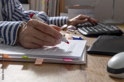 Closeup of woman hand who holding red marker and cheking text for grammar and using computer.Office table and documents,keyboard,mouse,tablet,pile of books on it