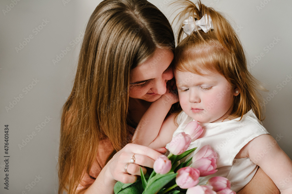 The mother holds on his hands little girl on light background indoor. Portrait of mom with flowers and daughter sitting in home. The concept of happy family holiday. woman's day. Mother's Day. The mother holds on his hands little girl on light background indoor. Portrait of mom with flowers and daughter sitting in home. The concept of happy family holiday. woman's day. Mother's Day.