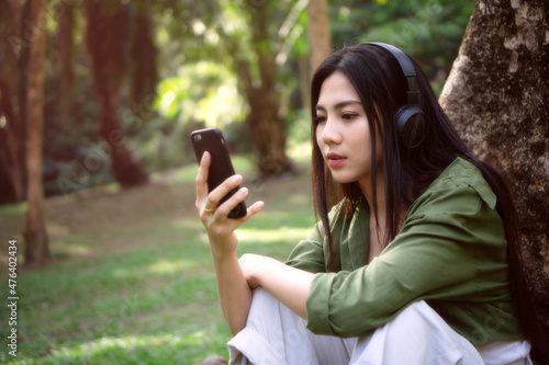 girl using headphone enjoy listening music under tree,lady sitting in outdoor park,nature as background,green jacket