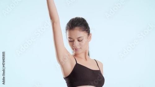 Horizontal medium shot of slim young pretty Asian brunette woman with long hair in ponytail in black bra strokes her raised up hand and armpit on pale blue background | After woman shaving concept photo