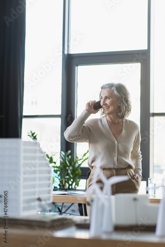 Positive businesswoman talking on cellphone near blurred models of buildings in office.