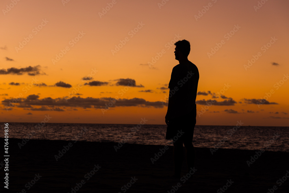 Man standing on a tropical beach at twilight silhouetted against a vivid orange ocean sunset