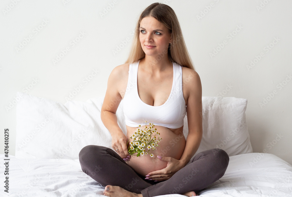 Cute blonde pregnant girl is sitting on the bed at home. Holds a flower twig near the belly. On a light background. Portrait. The concept of waiting for the birth of a child. Cute blonde pregnant girl is sitting on the bed at home. Holds a flower twig near the belly. On a light background. Portrait. The concept of waiting for the birth of a child.