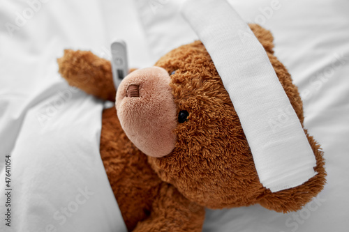 medicine, healthcare and childhood concept - ill teddy bear toy with cooling head bandage and thermometer lying in bed