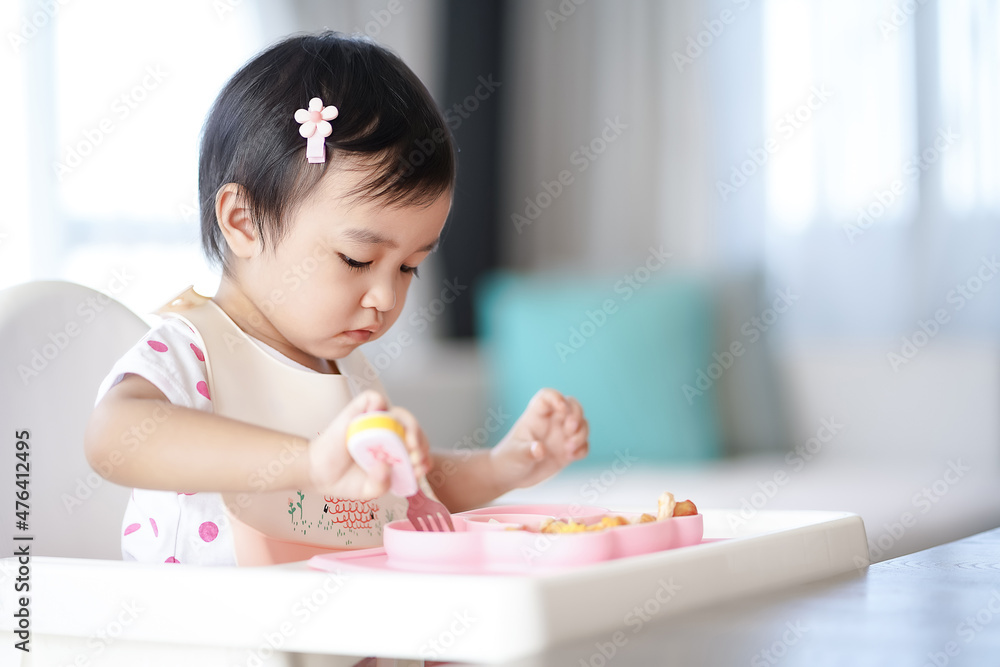 Asian cute little girl holding fork and trying to eat food sitting on the white high chair in the morning at home with copy space. Asian cute little girl holding fork and trying to eat food sitting on the white high chair in the morning at home with copy space.