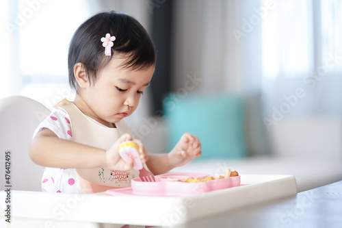 Asian cute little girl holding fork and trying to eat food sitting on the white high chair in the morning at home with copy space.