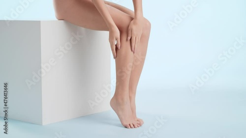 Horizontal shot of slim young attractive woman strokes her smooth leg sitting on the white platform on pale blue background | Moisturizer commercial photo