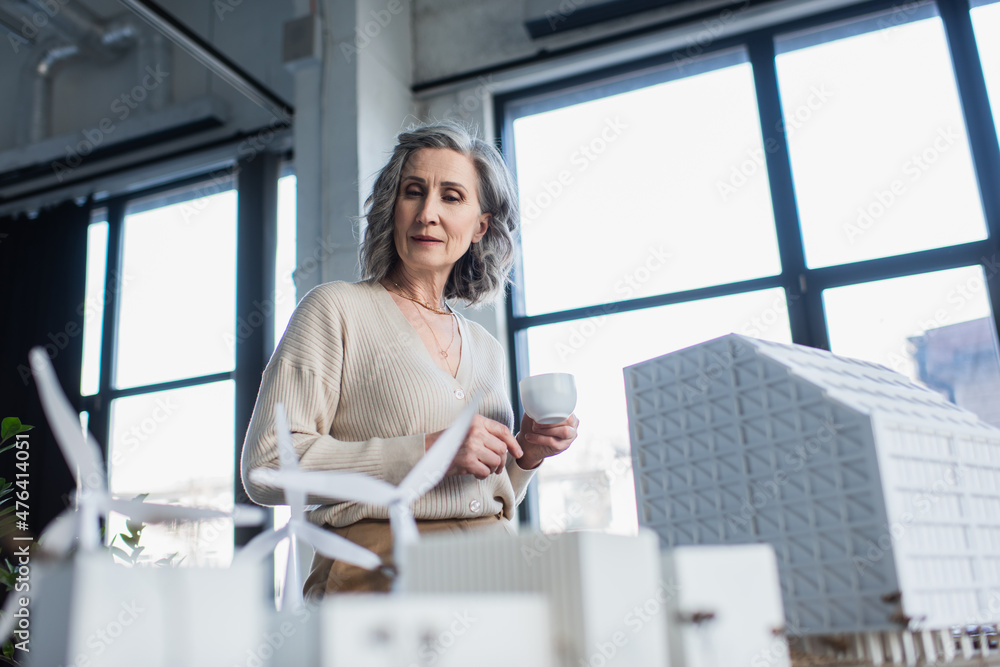 Grey haired businesswoman holding coffee cup and looking at models of buildings in office.