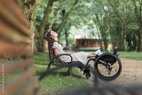 Caucasian disabled woman resting on wooden bench outdoors with feet on wheelchair. Pretty young woman looking up and enjoying every moment of her life. © sofiko14