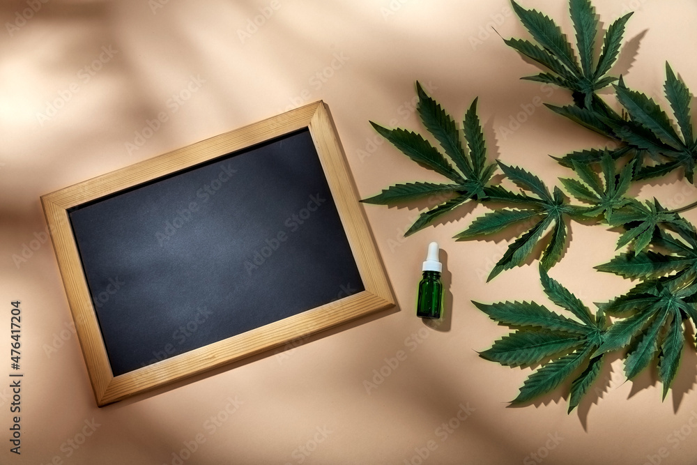 herbal medicine and aroma therapy concept - close up of hemp essential oil in glass bottle, chalkboard and cannabis leaves on beige background