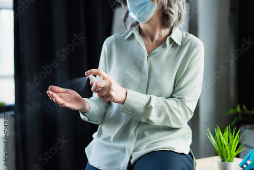 Cropped view of grey haired businesswoman in protective mask spraying hand sanitizer in office.