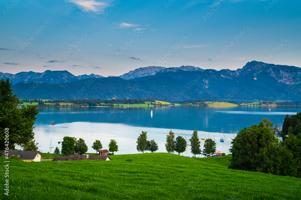 Germany, Allgaeu, Boats and sailing ships on silent water of forgensee lake with spectacular alps panorama at sunset in summer atmosphere