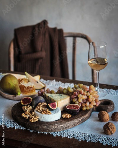 Appetizer. A glass of white wine, grapes, figs, pear, nuts and honey on a black wooden board. Dor blue cheese, brie cheese on a dark table. Sweater on a chair. Background image, copy space. Autumn