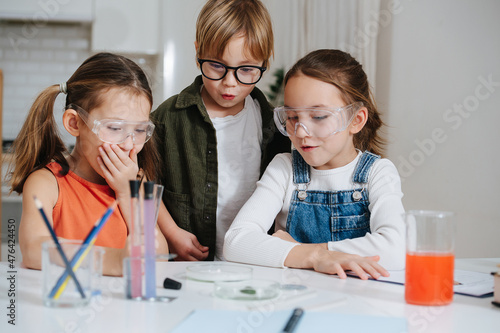 Astonished little kids doing home science project, looking at the glass dish in amasment. All wearing glasses. Chemical glassware and colored liquids on the table.
