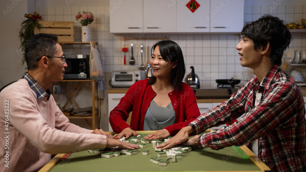 happy mother chatting with father and son while they are playing mahjong together in the evening during spring festival. text on cabinet translation: luck