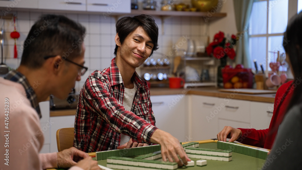 selective focus of smiling asian son looking and keeping the tile he picks and taking out one from his hand while playing mahjong at home in the evening during spring festival