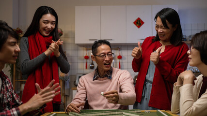 excited mother and daughter cheering and massaging the father as he wins by his own draw on mahjong gambling table. chinese text at background translation: luck