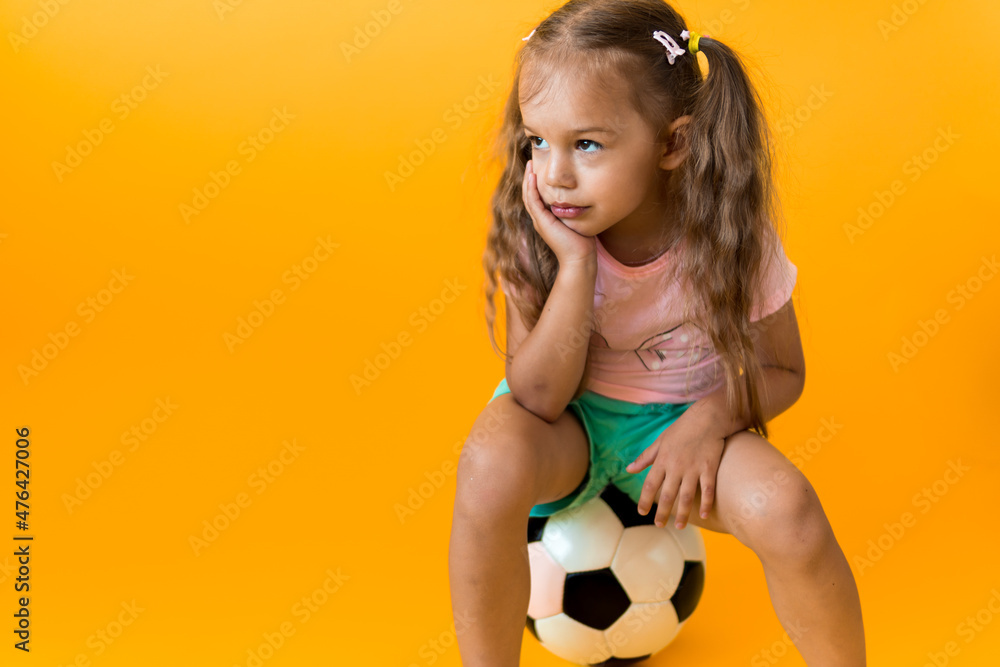 Authentic cute smiling preschool little girl with classic black and white soccer ball look at camera on yellow background. child play football in t-shirt and shorts. Sport, championship, team concept Authentic cute smiling preschool little girl with classic black and white soccer ball look at camera on yellow background. child play football in t-shirt and shorts. Sport, championship, team concept